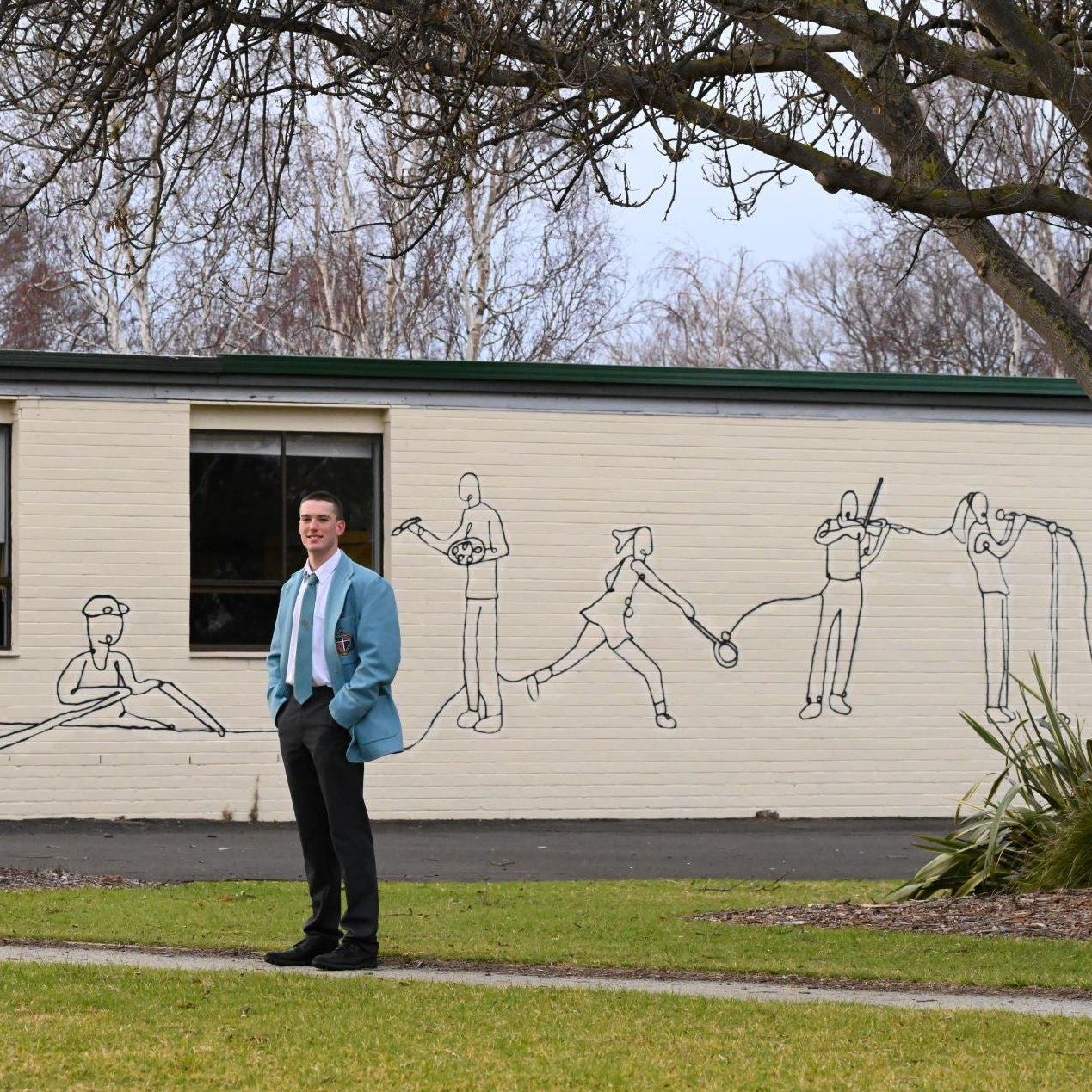 Man standing in front of a building with a mural of children playing sports on the wall.