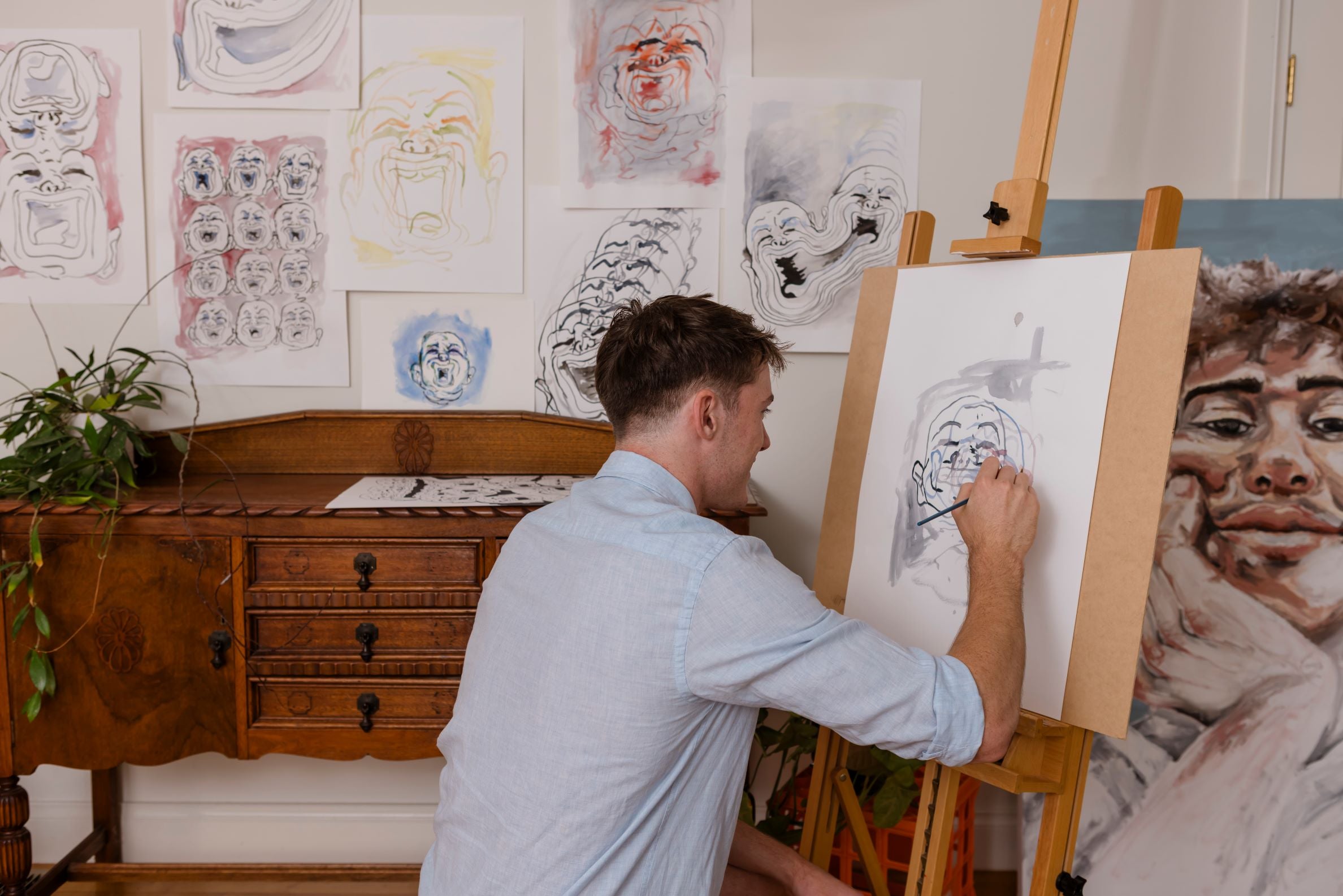 Man painting a portrait in an artist's studio with various artworks on the walls.